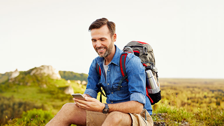 Lächelnder Mann beim Wandern macht eine Pause und schaut auf sein Handy