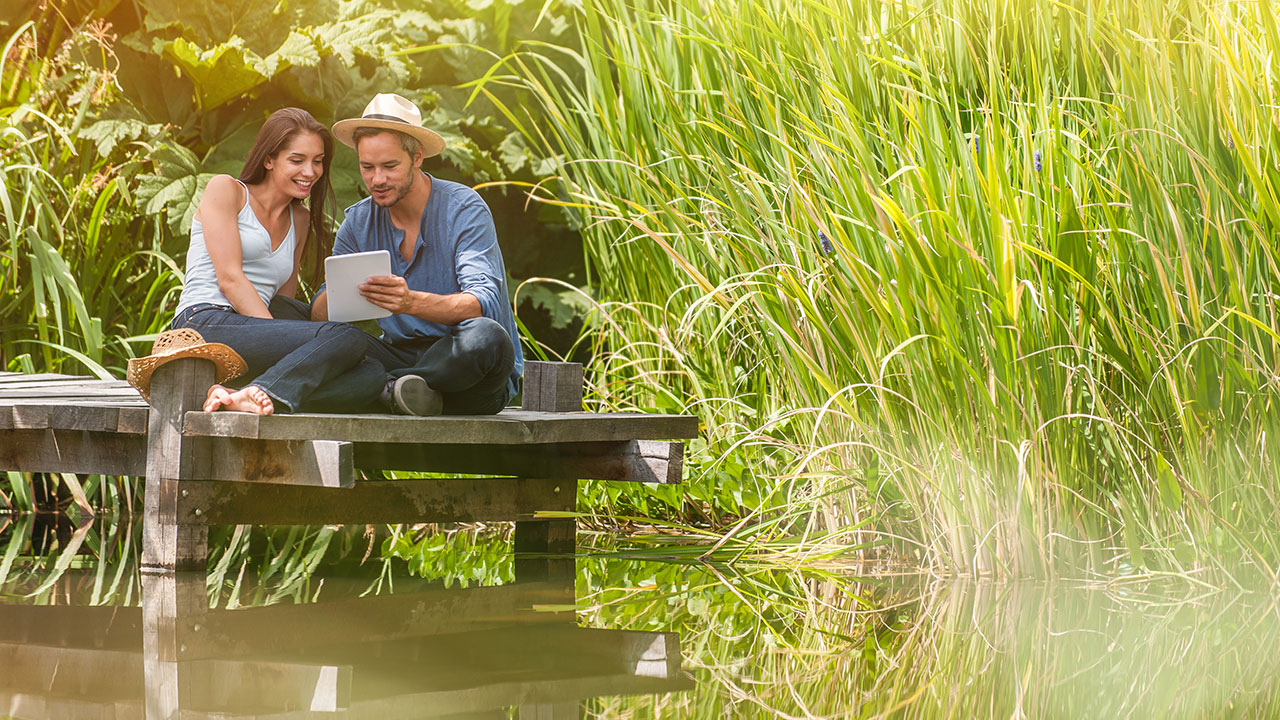 Tagesgeld auf einen Blick Pärchen sitzt entspannt am Steg und blickt auf ein Tablet