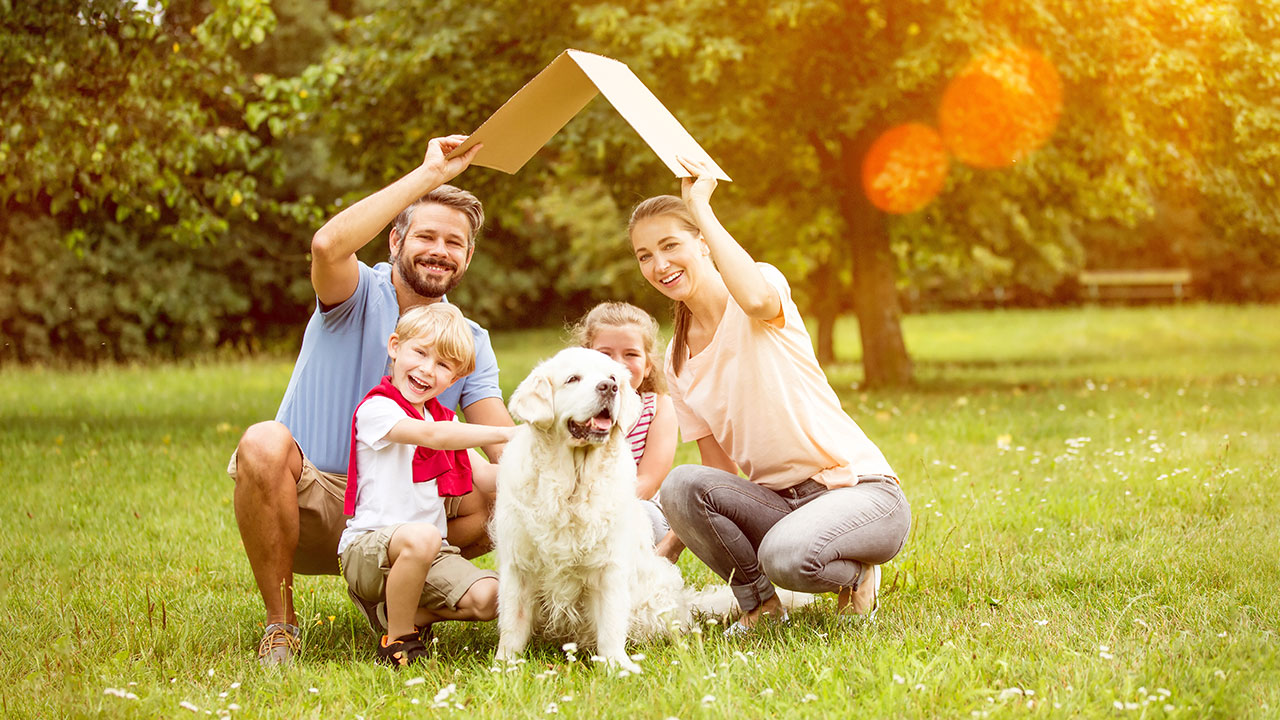Sparziel - Eigenheim Vierköpfige Familie mit Hund hält ein Kartondach über Ihren Kopf als Symbol für ein Eigenheim